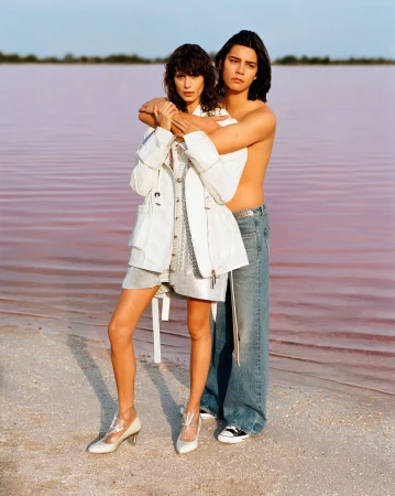 Model wearing a white jacket over a patterned dress and heels, embraced by a shirtless man in denim jeans standing on a sandy shore by water