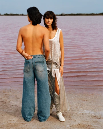 A man in denim jeans and a woman in a silver dress stand together by a pink body of water for the Rabanne Spring 2025 campaign.