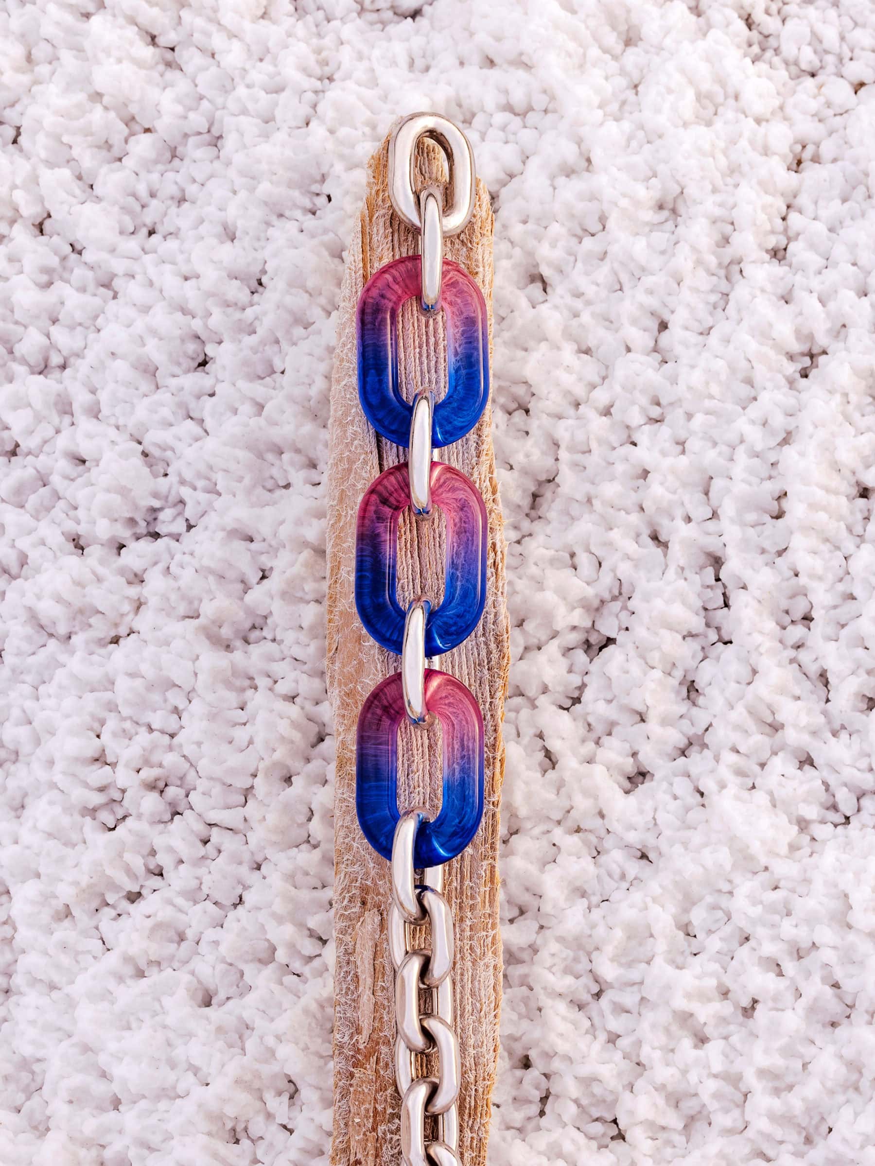 Close-up vertical shot of a metallic and blue-red resin chain link necklace resting on textured sand.