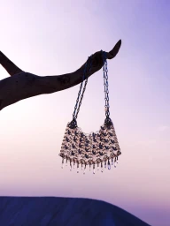 Metallic mesh handbag hanging from a dark tree branch against a sunset sky and dark landscape background