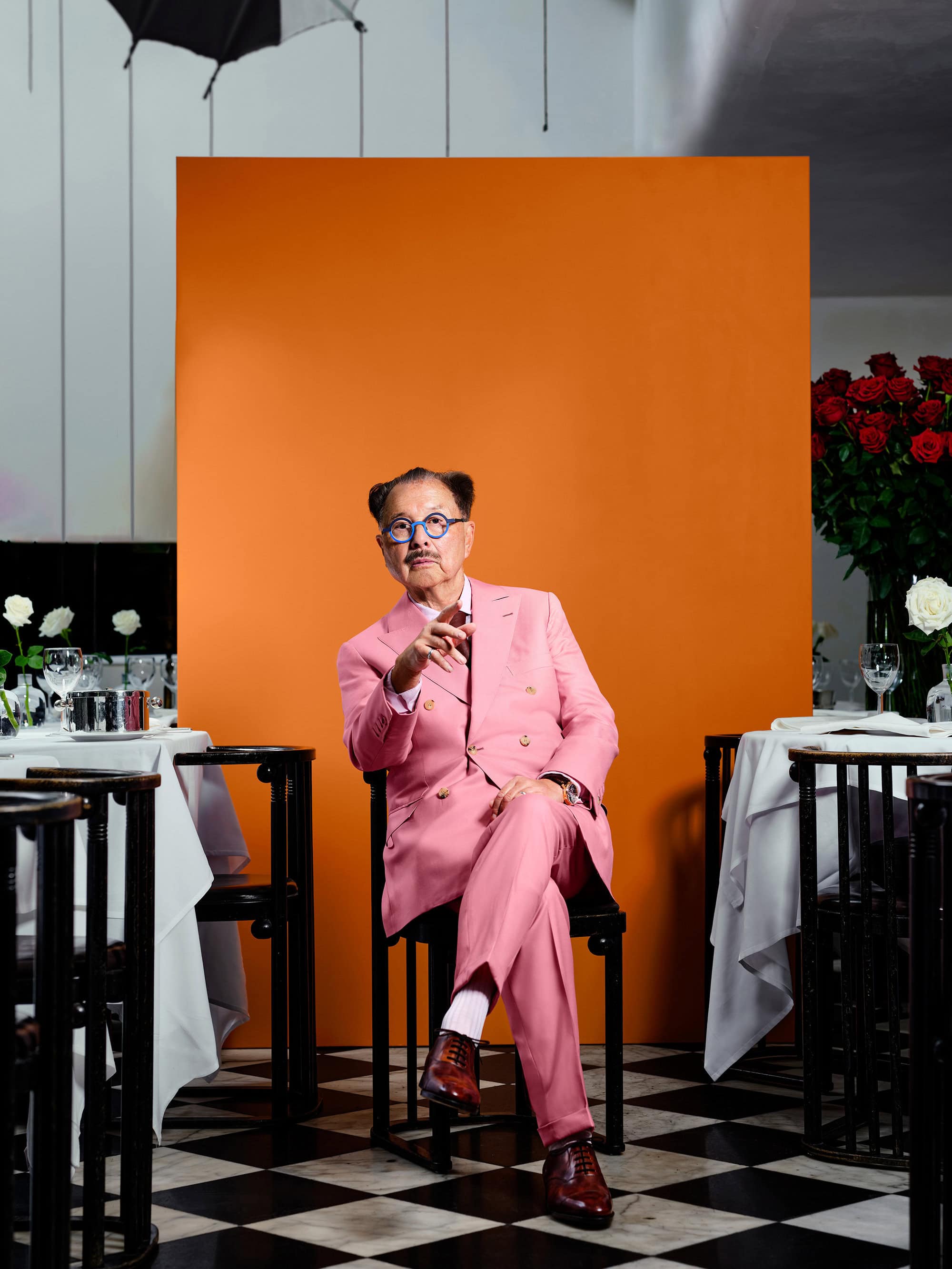 Man wearing a pink suit and blue glasses seated on a chair against a vibrant orange backdrop in a restaurant.