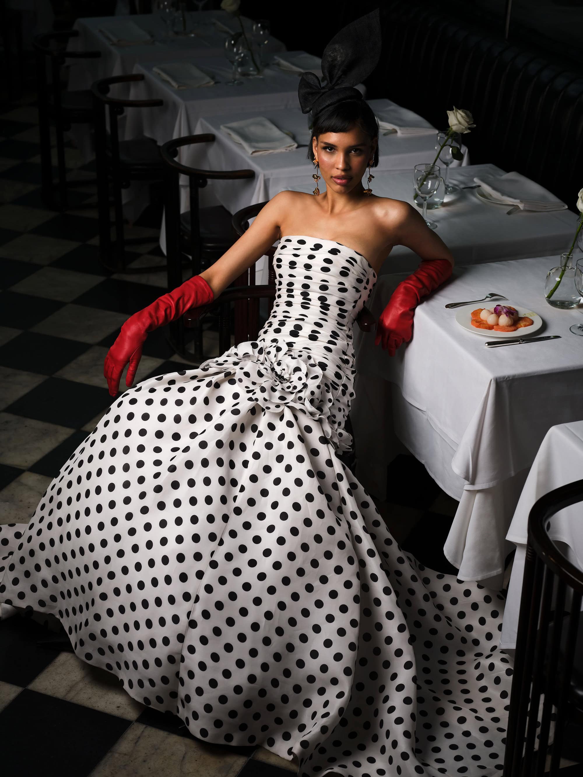 Model in polkadot gown and red gloves seated at a restaurant table for Neiman Marcus