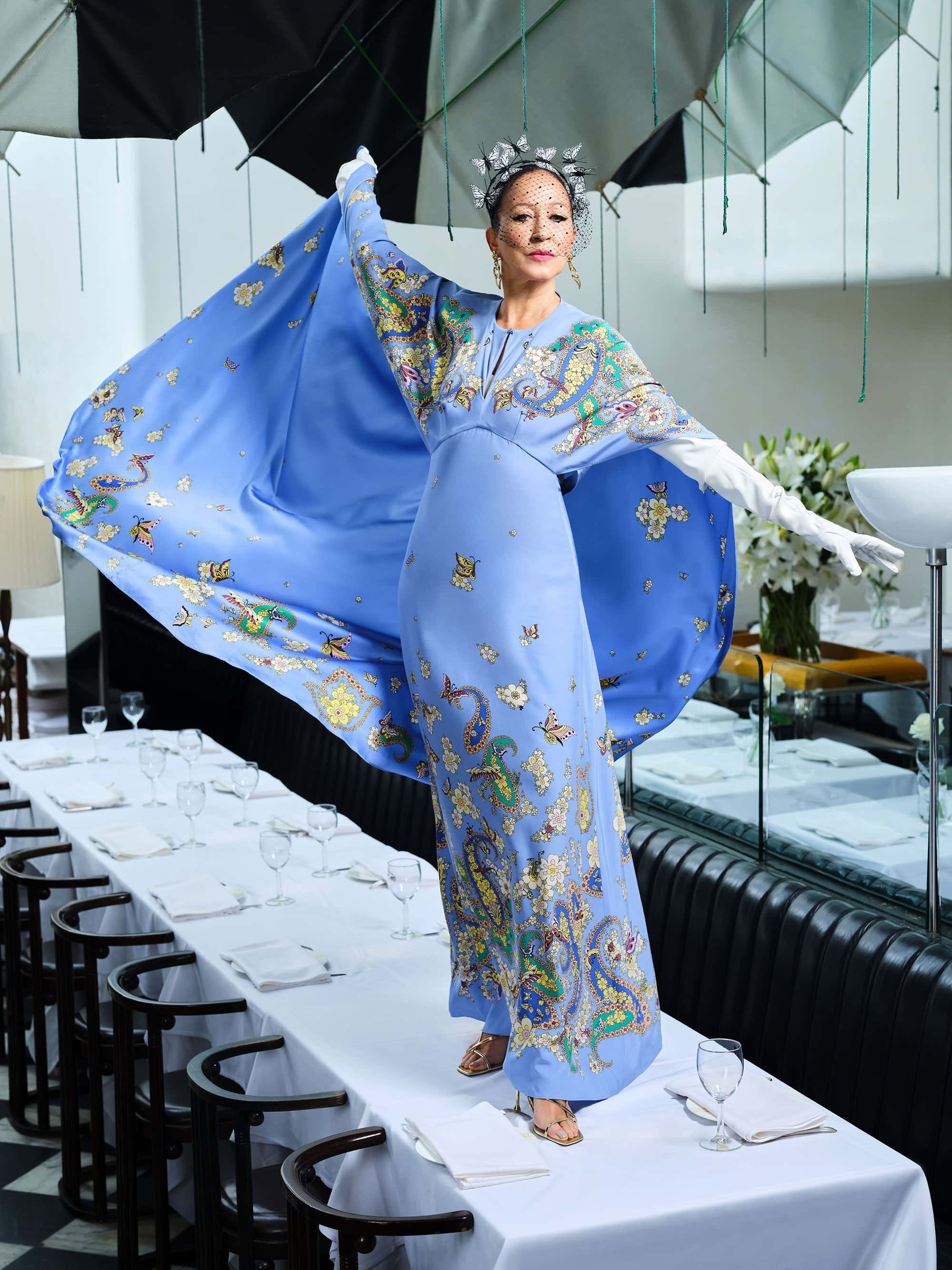 Woman in blue patterned gown with a dramatic cape standing on a restaurant dining table under hanging umbrellas