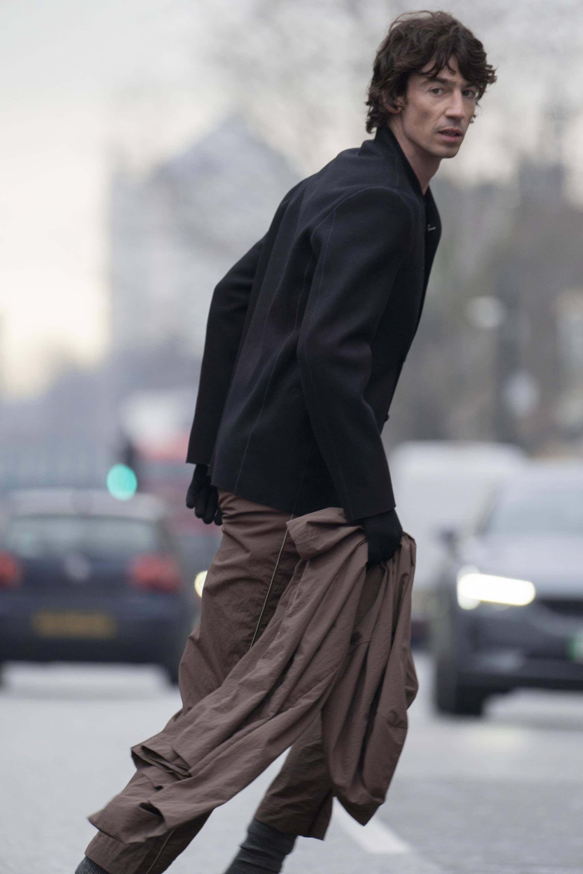 Man wearing a black jacket and brown trousers crossing an urban street, holding an extra garment.