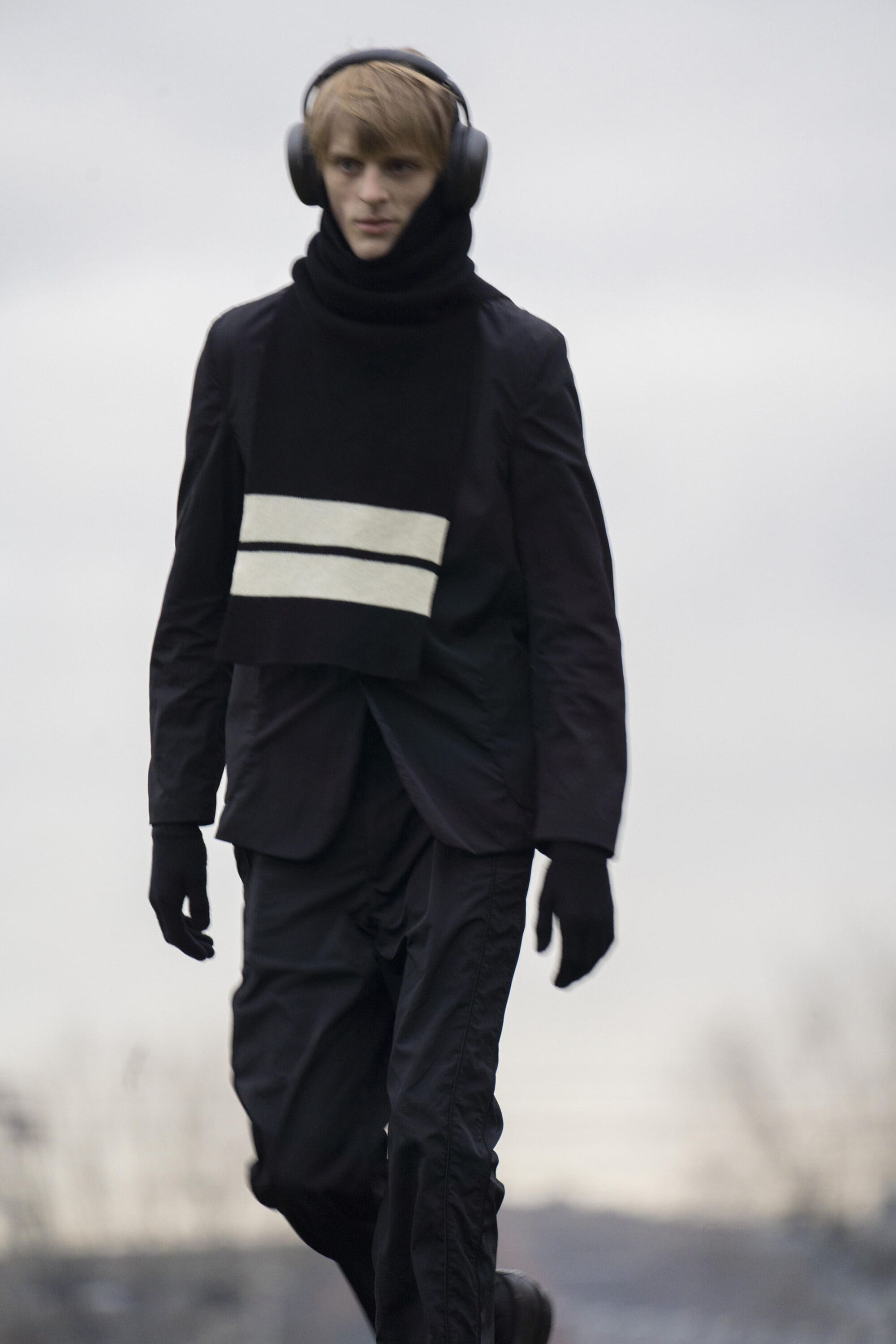 A young male model wearing black winter outerwear and over-ear headphones walking outdoors under an overcast sky.