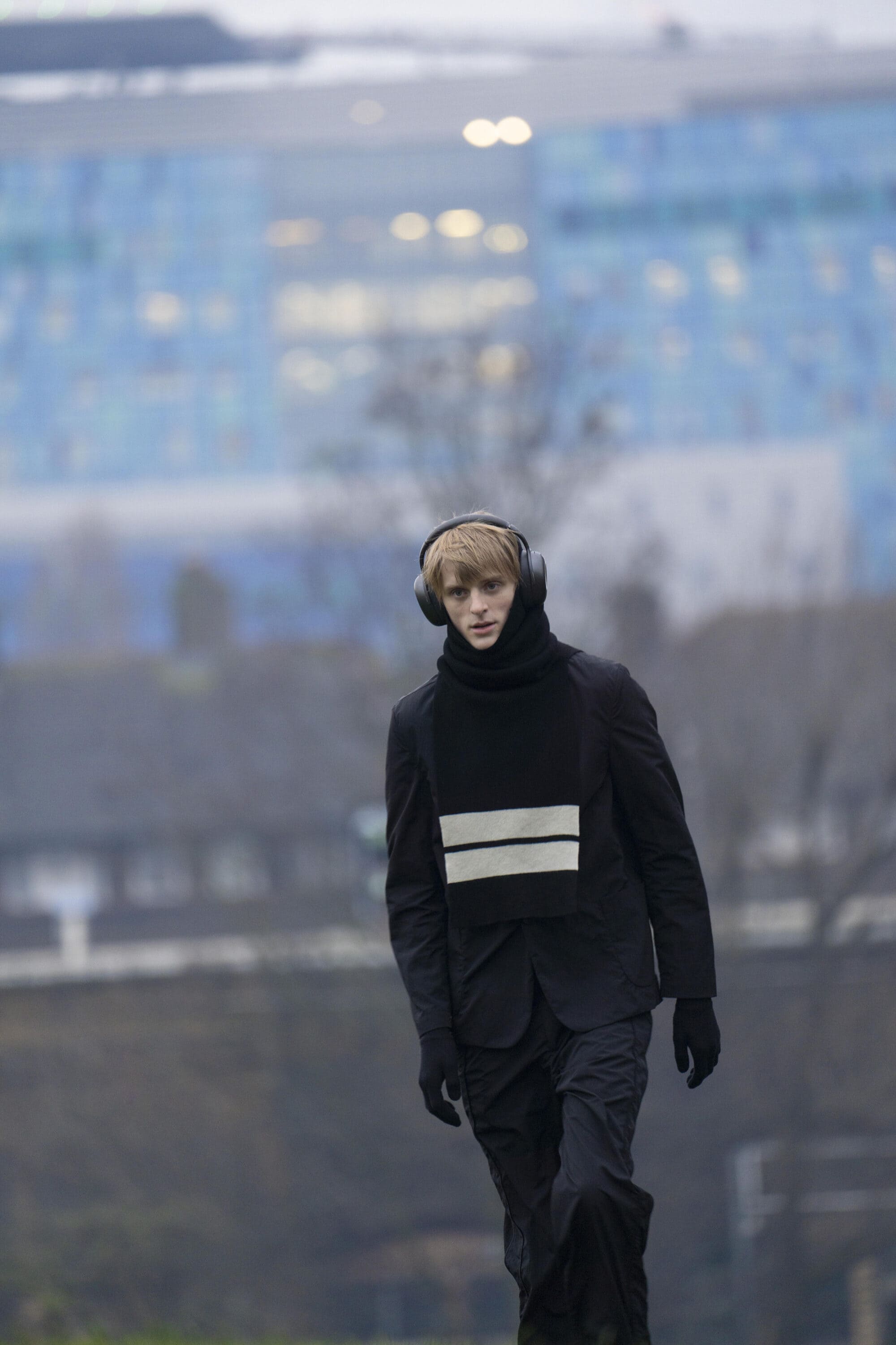 A young male model walking outdoors wearing a black jacket and striped scarf with headphones against a blurred industrial background.