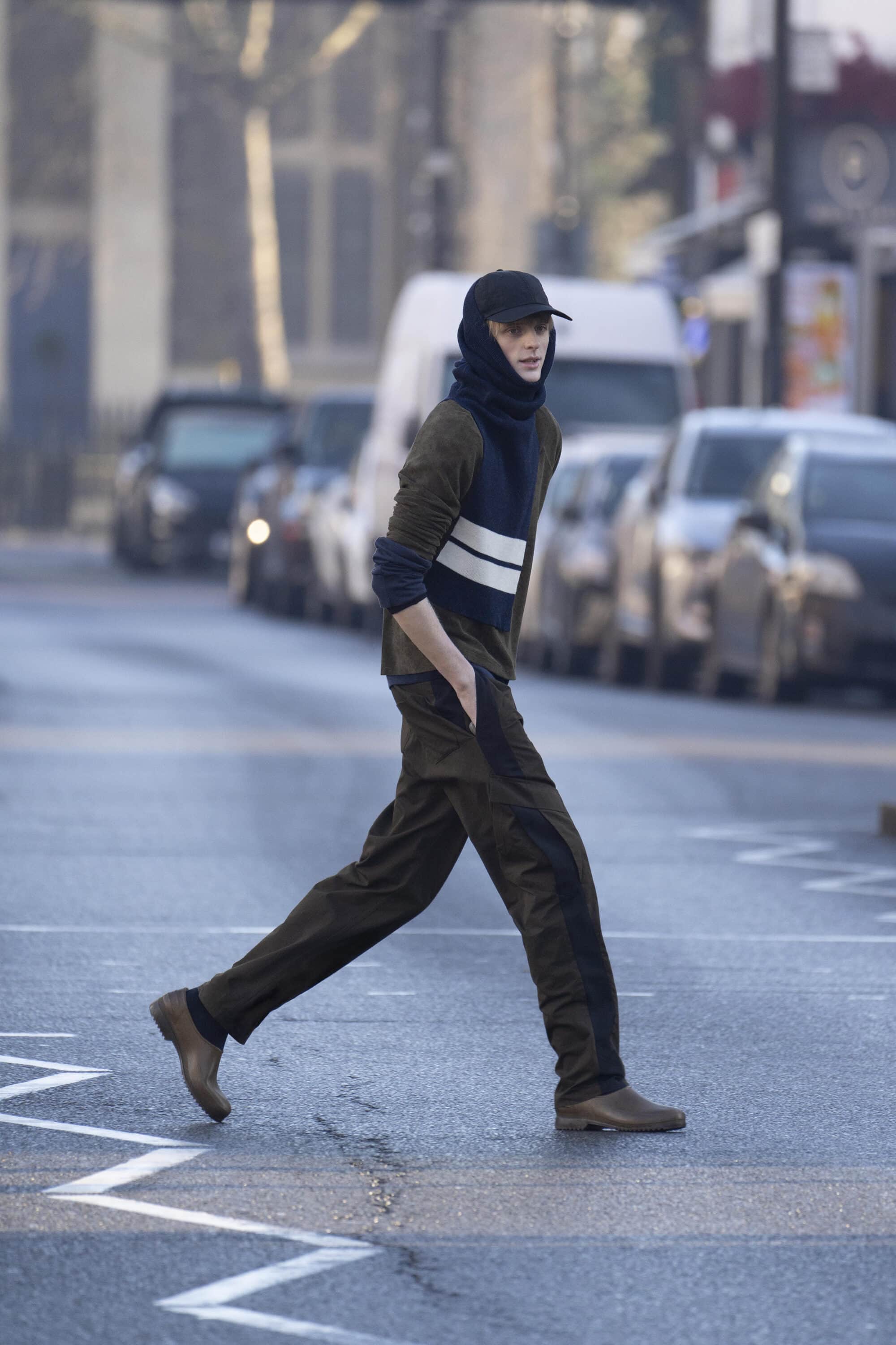 Model wearing dark trousers and baseball cap walking across a city street for the Derrick Intermission campaign