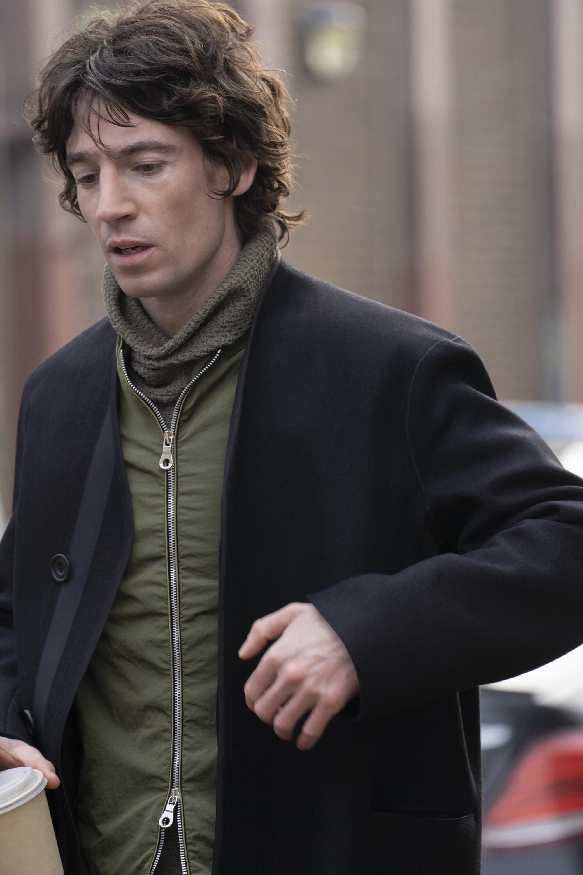 Man wearing a black coat and olive green zippered jacket walking outdoors for Derrick