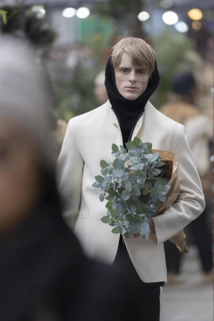 Young man wearing a cream jacket and black hood holding a bouquet of eucalyptus in a market