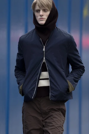 Model wearing dark blue zip-up bomber jacket and brown trousers with head wrapped in dark cowl for Derrick Intermission campaign
