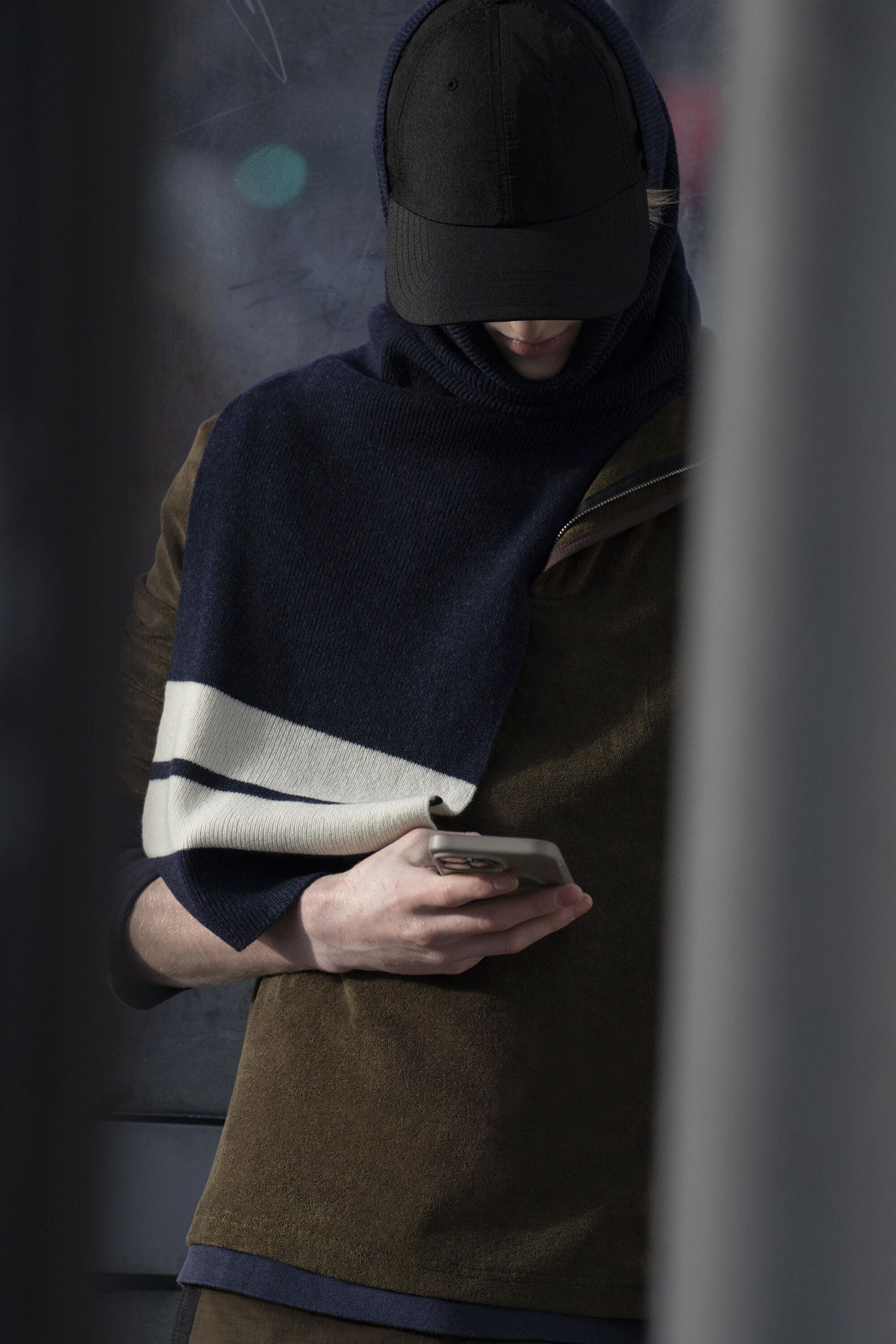 Man wearing a dark baseball cap, navy scarf, and brown top holding a smartphone