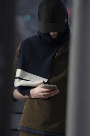 Man wearing a dark baseball cap, navy scarf, and brown top holding a smartphone