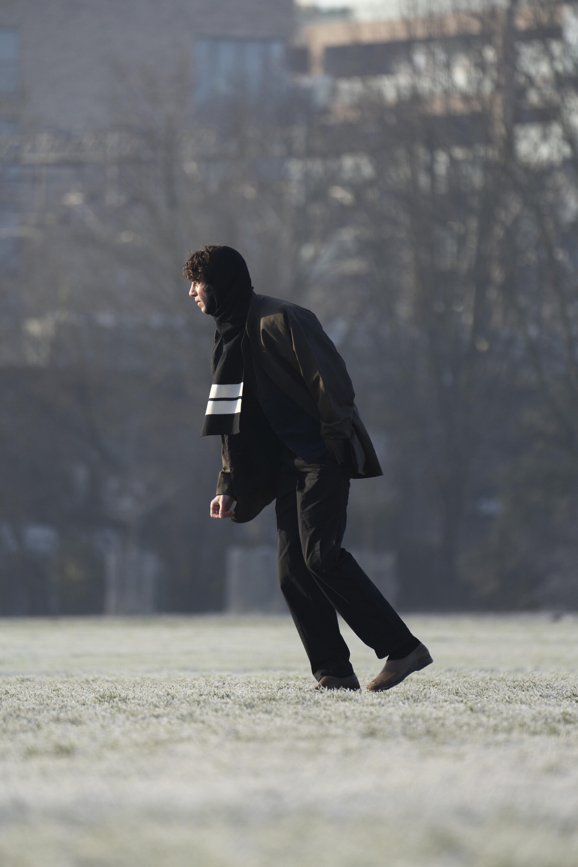 Man walking across a frosty field wearing a dark jacket, black trousers, and a scarf with white stripes