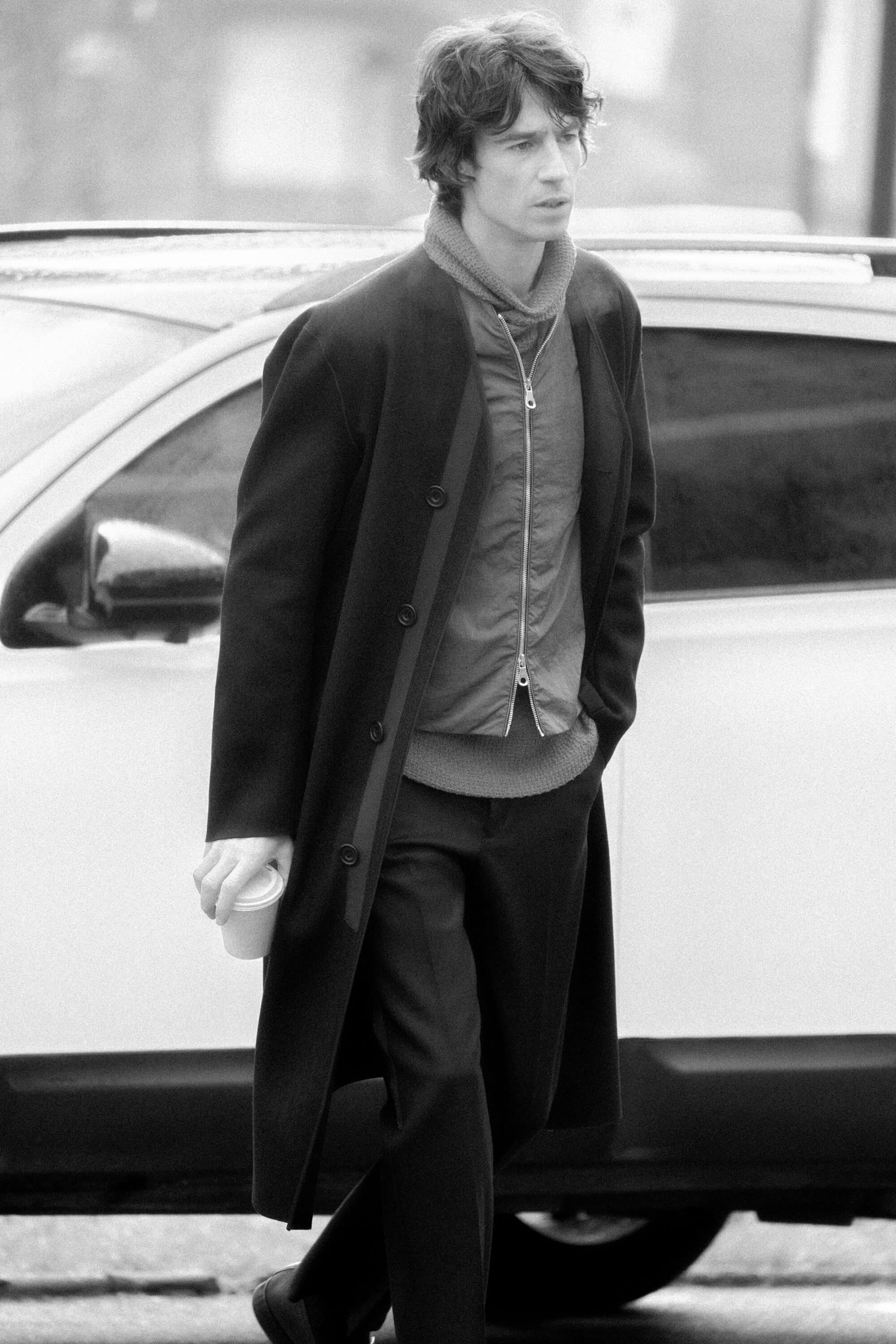 Man wearing a long black coat and trousers walking past a car holding a cup
