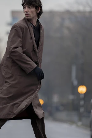 Man wearing a long brown coat and black gloves walking outdoors against a blurred city street background.