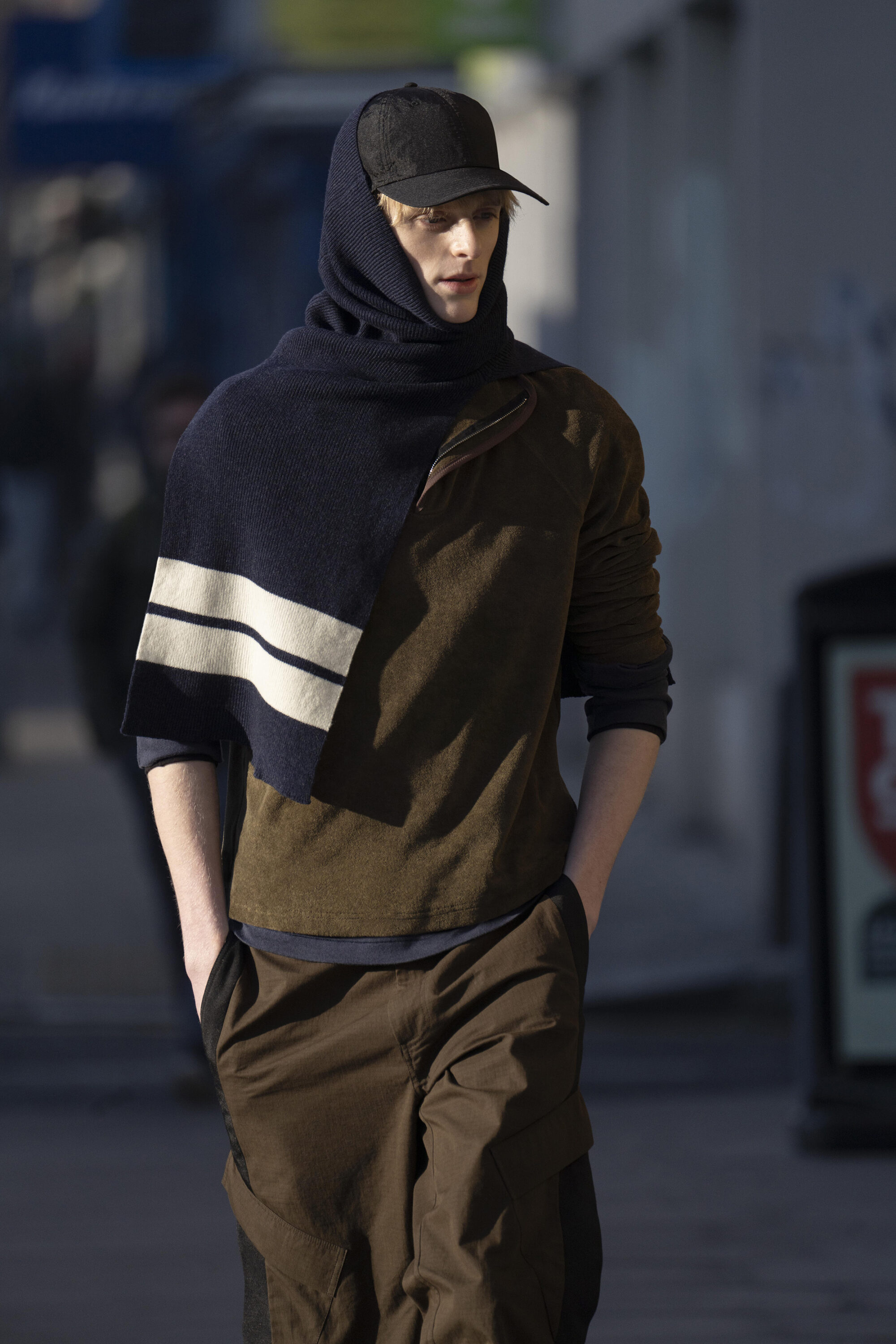 Model wearing a dark cap, hooded dark scarf with stripes, olive top, and brown trousers walking outdoors for Derrick
