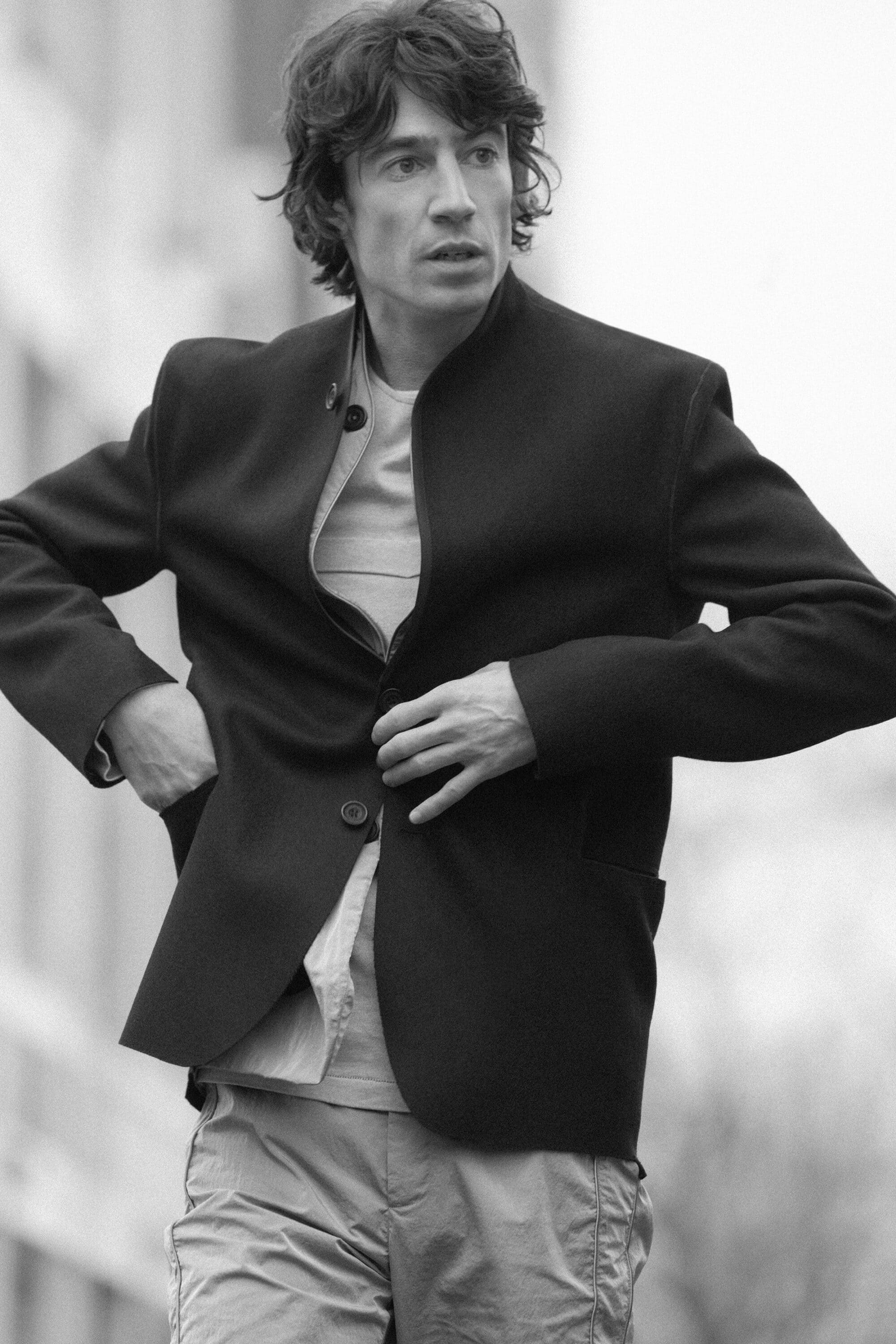 Black and white portrait of a man wearing a dark jacket over a t-shirt adjusting his button in an outdoor setting