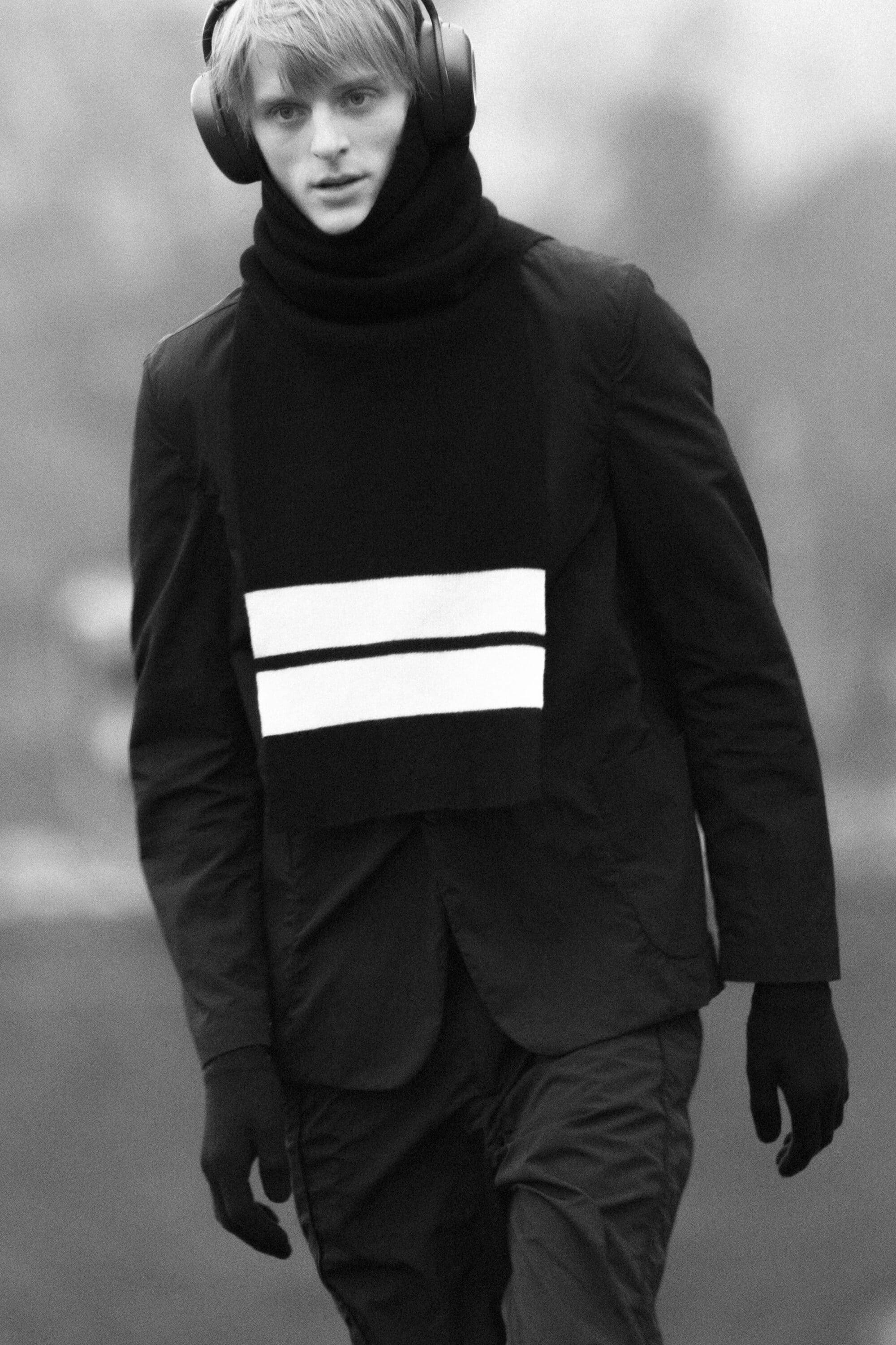 Young male model in a black jacket and striped scarf wearing headphones outdoors
