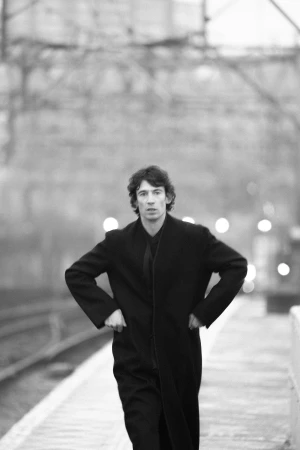 A man in a black coat standing on a railway platform with hands on his hips in a black and white portrait.