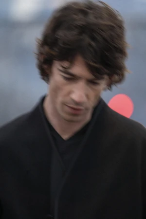 Portrait of a man wearing a black coat with dark hair looking downward outdoors