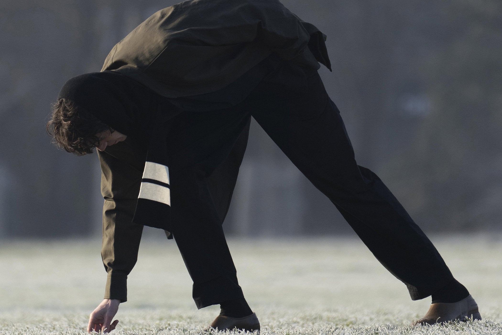 Model bending forward in a dark hooded jacket and trousers on a frosty field