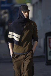 Model wearing a dark cap, hooded dark scarf with stripes, olive top, and brown trousers walking outdoors for Derrick