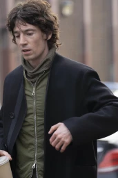 Man wearing a black coat and olive green zippered jacket walking outdoors for Derrick