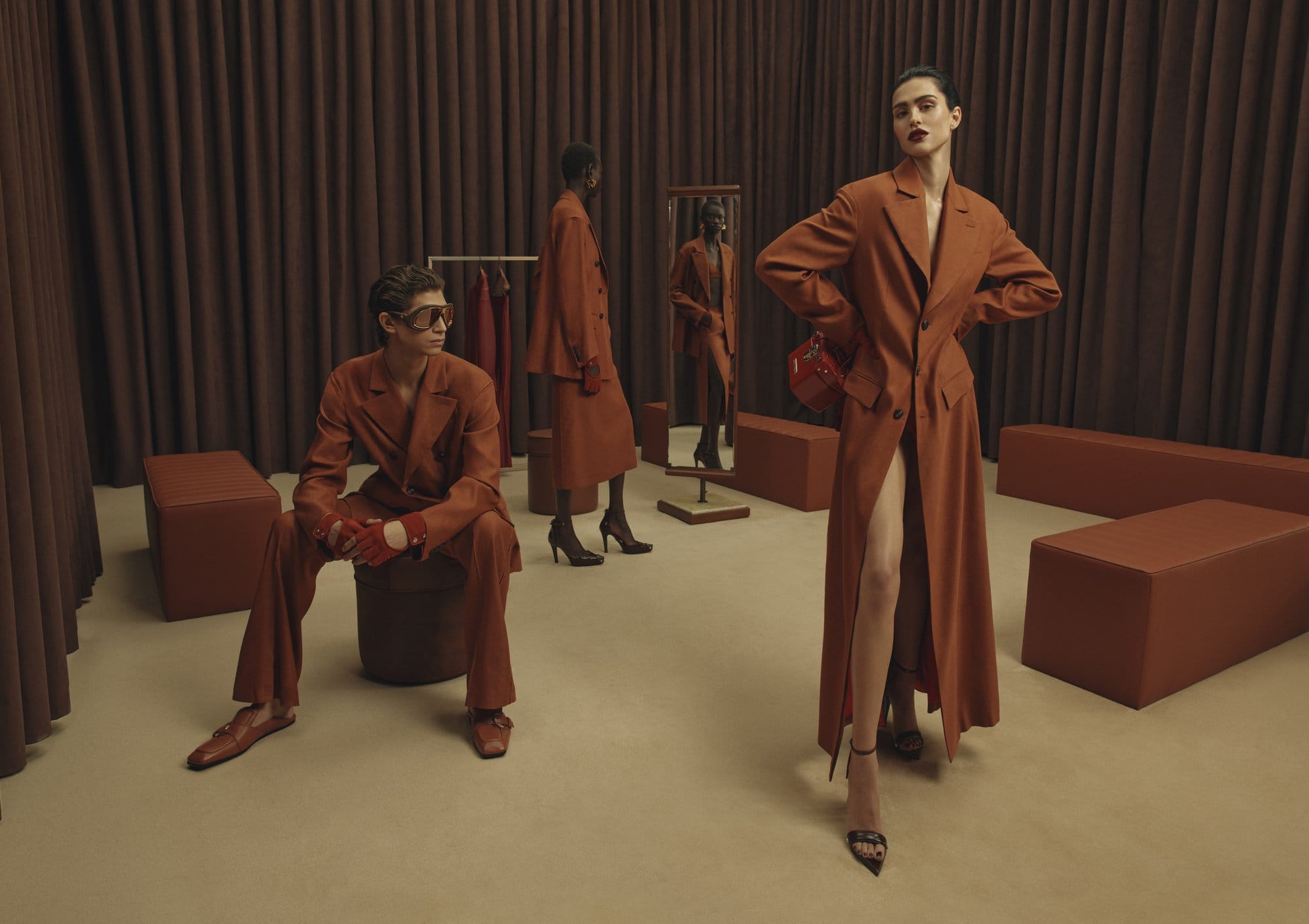 Three models wearing rust-colored tailored suits inside a showroom with brown curtains and matching furniture.