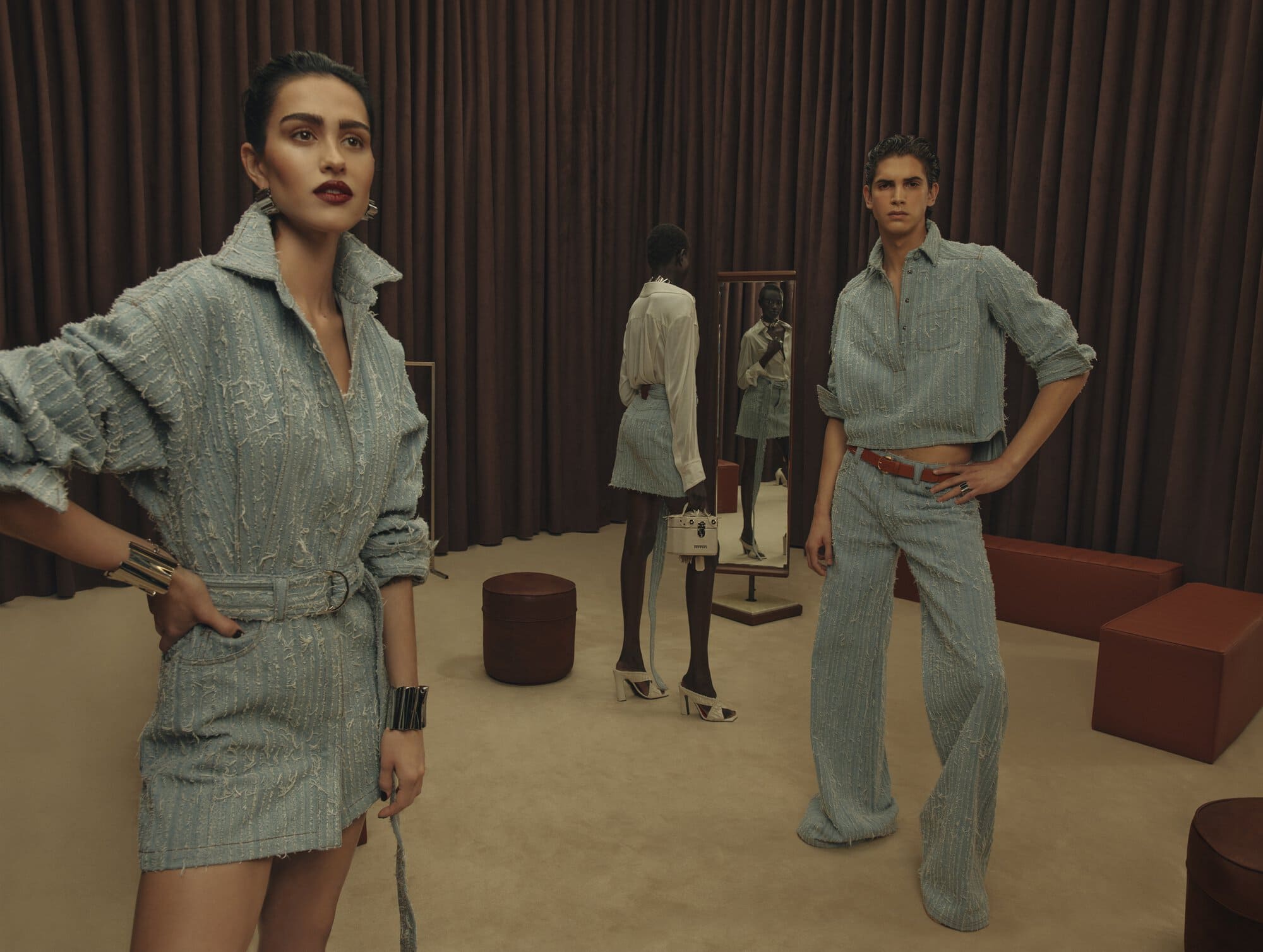 Models in textured light blue denim clothing posing in a studio with dark brown curtains and leather ottomans for Ferrari Spring 2025.