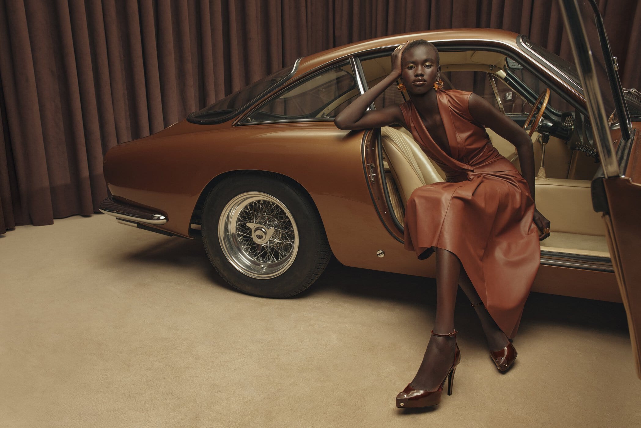 Model seated inside a classic bronze car wearing a brown leather dress against a warm studio background.