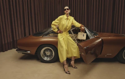 Model in a yellow dress and sunglasses posing next to a brown classic car in front of brown curtains