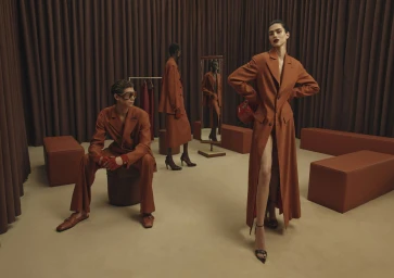 Three models wearing rust-colored tailored suits inside a showroom with brown curtains and matching furniture.