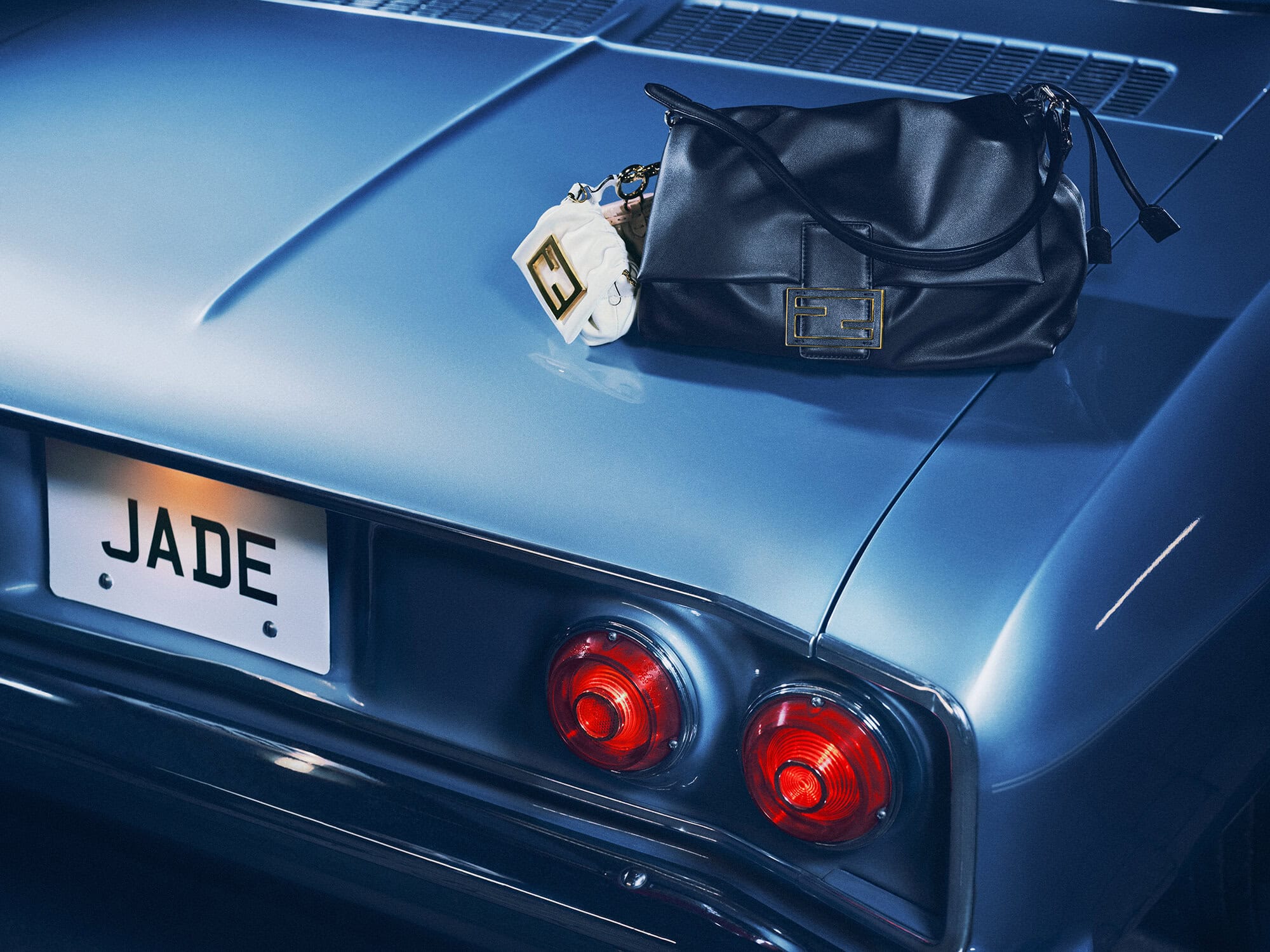 Black leather Fendi Baguette handbag resting on the rear trunk of a vintage blue car with a custom JADE license plate.
