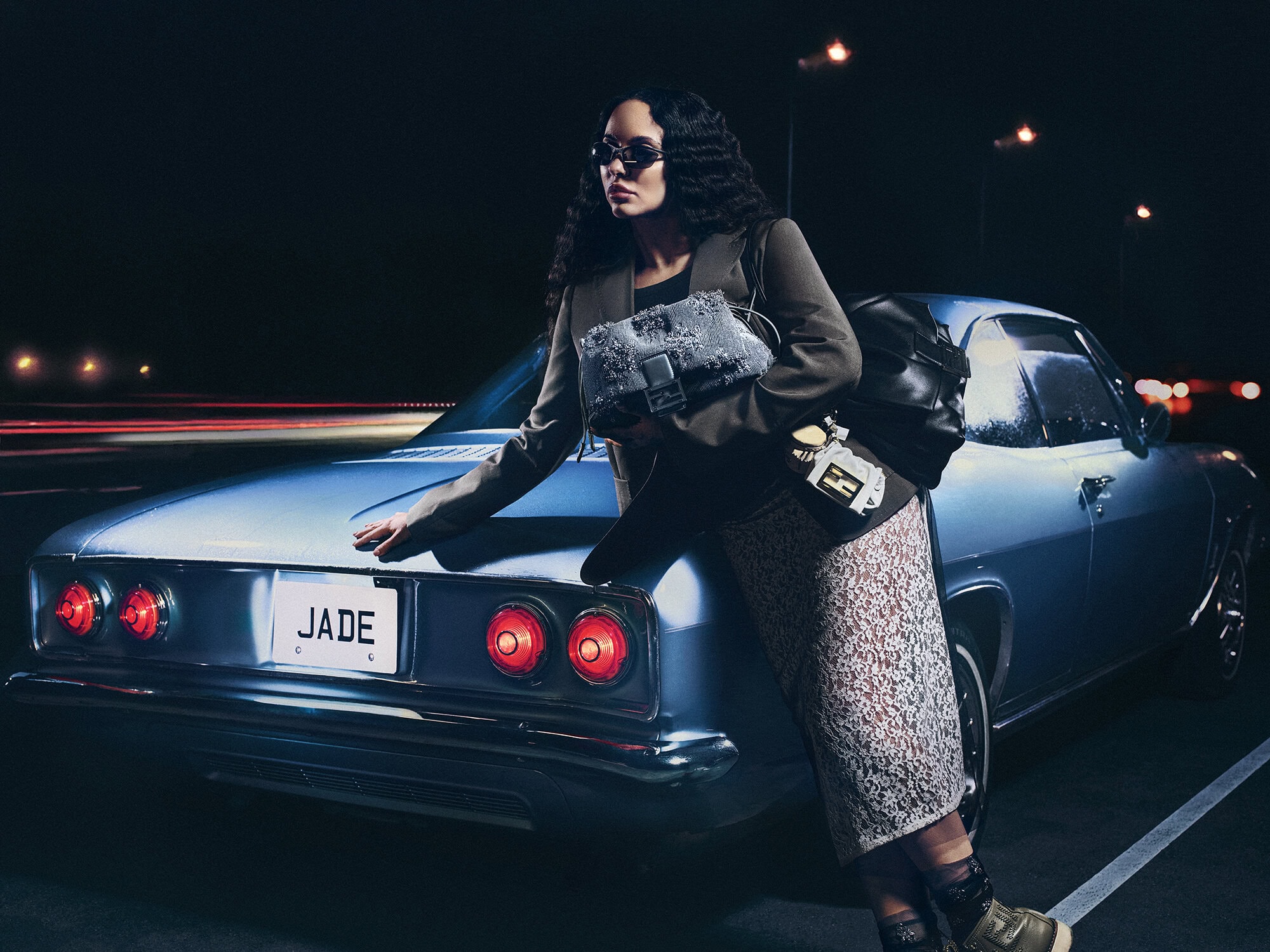 Jade Thirlwall leaning against a vintage blue car holding a Fendi Baguette bag at night under streetlights
