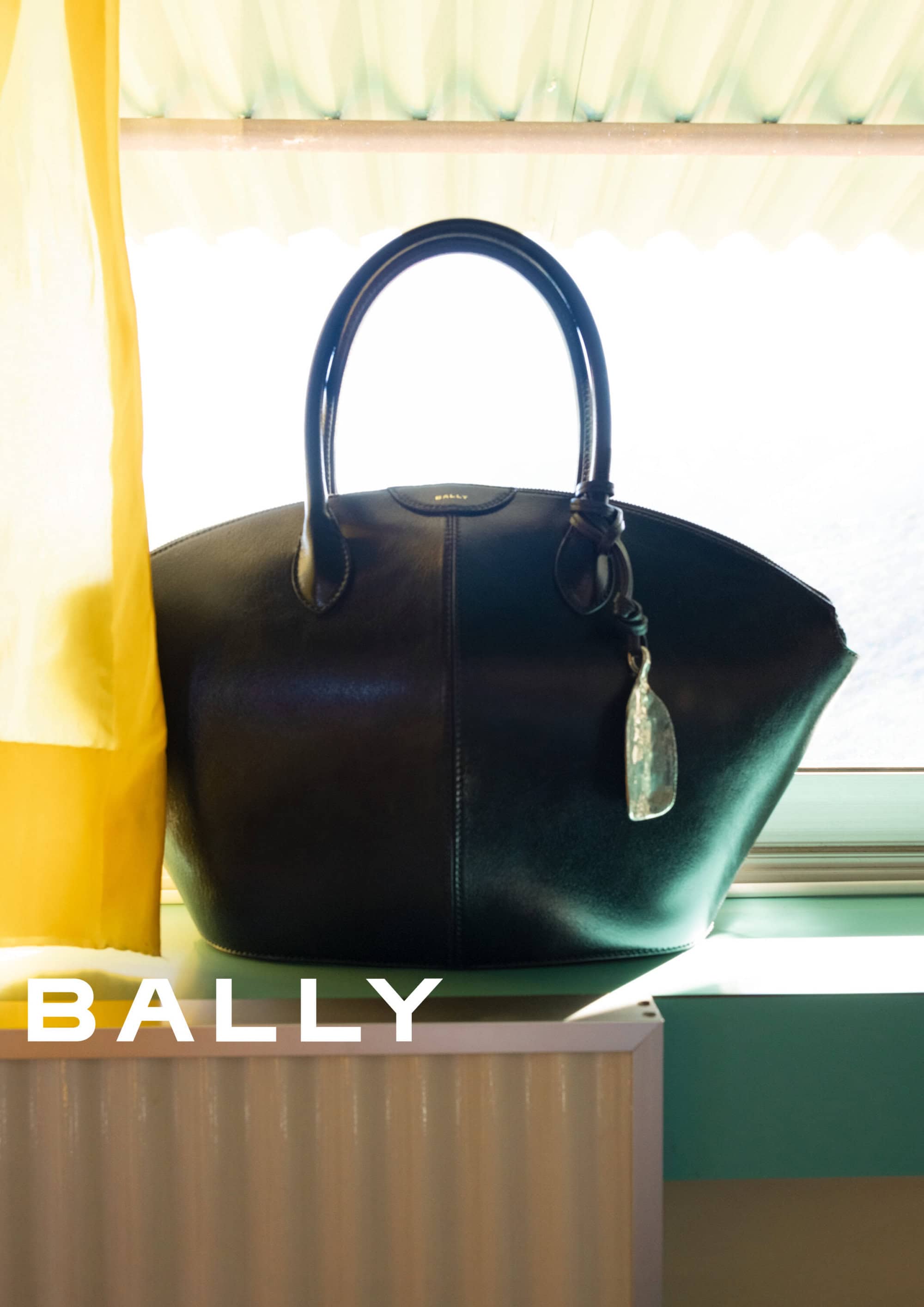 A black leather handbag resting on a window ledge beside a yellow curtain for Bally Spring 2025 ad campaign.