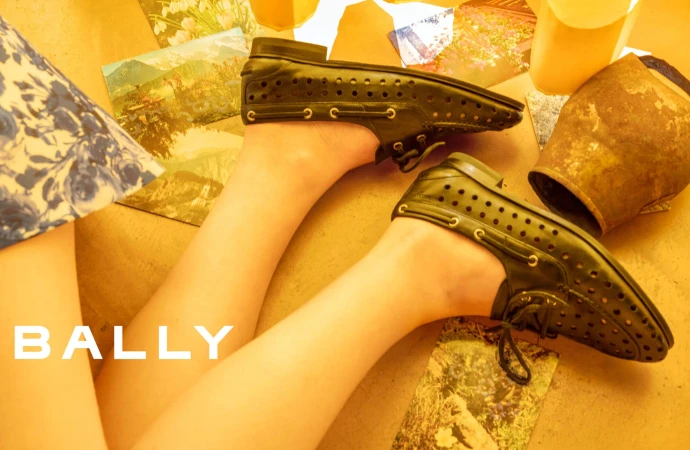 Close-up of perforated black leather loafers on legs against a warm yellow-toned flat lay background with vintage postcards.