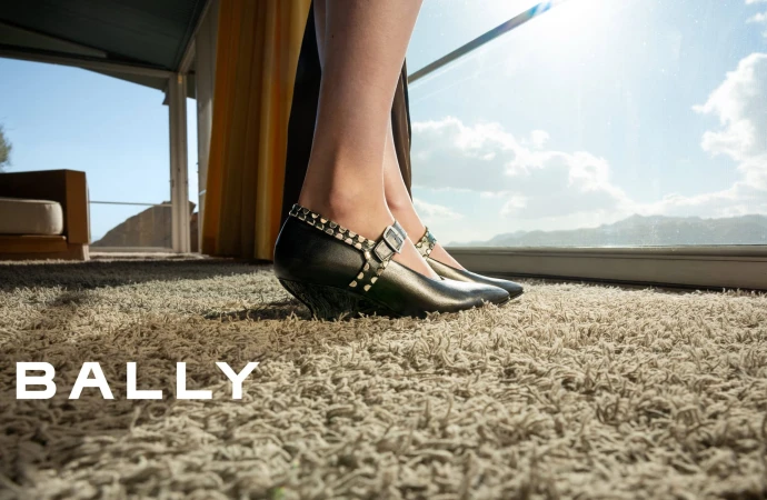 Close-up of a model's feet wearing buckled black shoes standing on a thick carpet inside a bright room by large windows.