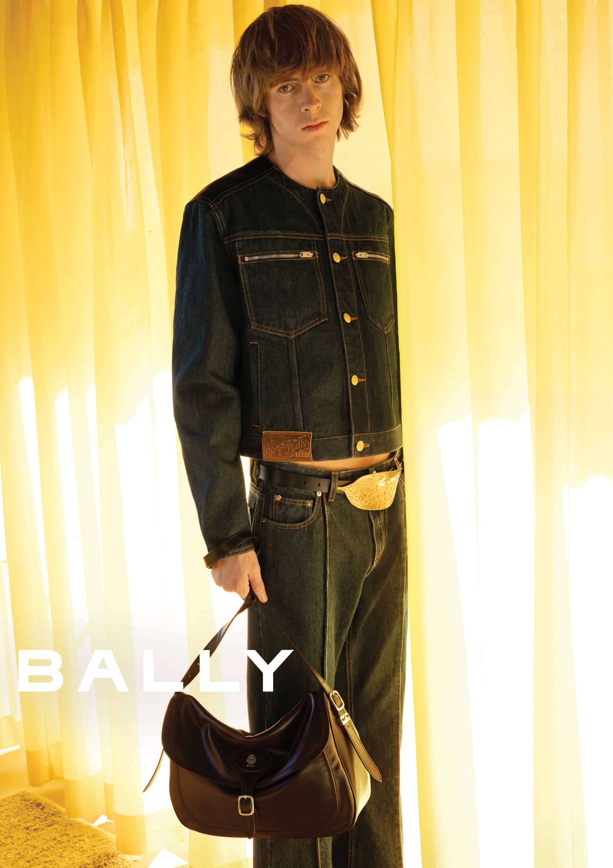 A male model in a dark denim jacket and matching jeans holding a brown leather shoulder bag against yellow curtains.