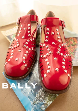 Close-up product shot of shiny red leather T-bar shoes with silver studs resting on open travel magazines