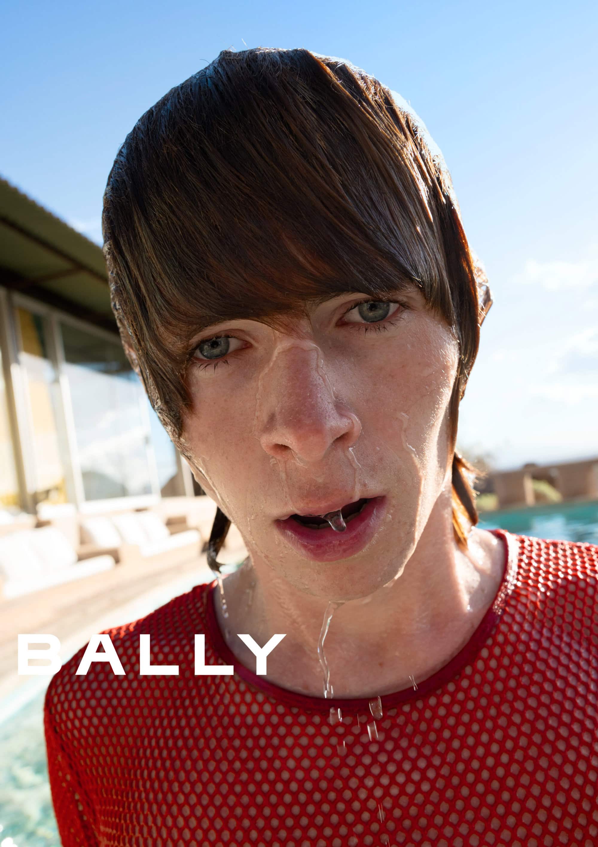 Close-up portrait of a young man with wet hair wearing a red mesh shirt outdoors near a pool for the Bally Spring 2025 ad campaign.