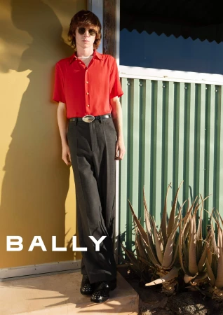 A person standing against a yellow and green building exterior wearing a red short-sleeve shirt and dark trousers for Bally.