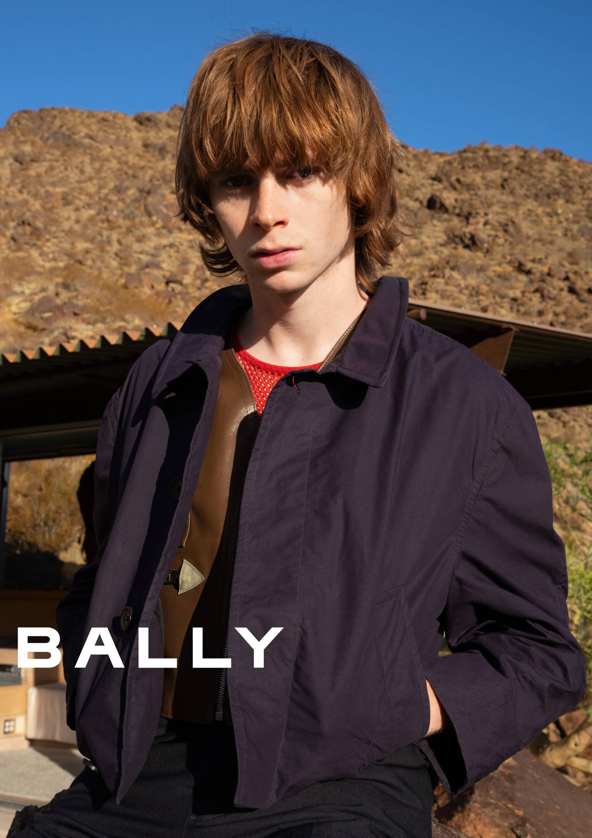 Young man with medium brown hair wearing a dark jacket outdoors against a rocky desert landscape for Bally Spring 2025 campaign.