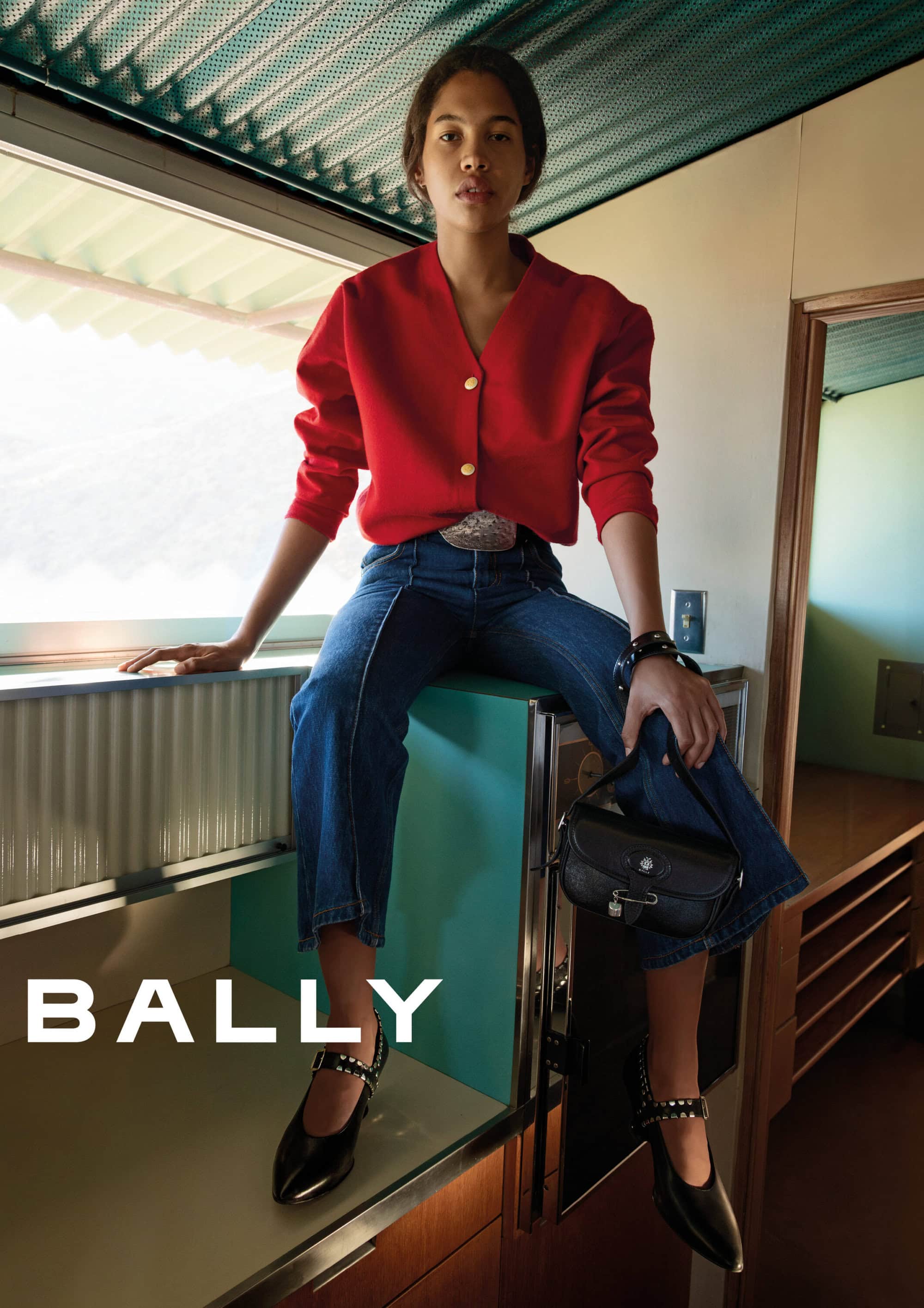 Model wearing a red cardigan and blue jeans sitting indoors holding a black leather handbag for Bally Spring 2025 campaign.