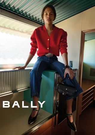 Model wearing a red cardigan and blue jeans sitting indoors holding a black leather handbag for Bally Spring 2025 campaign.