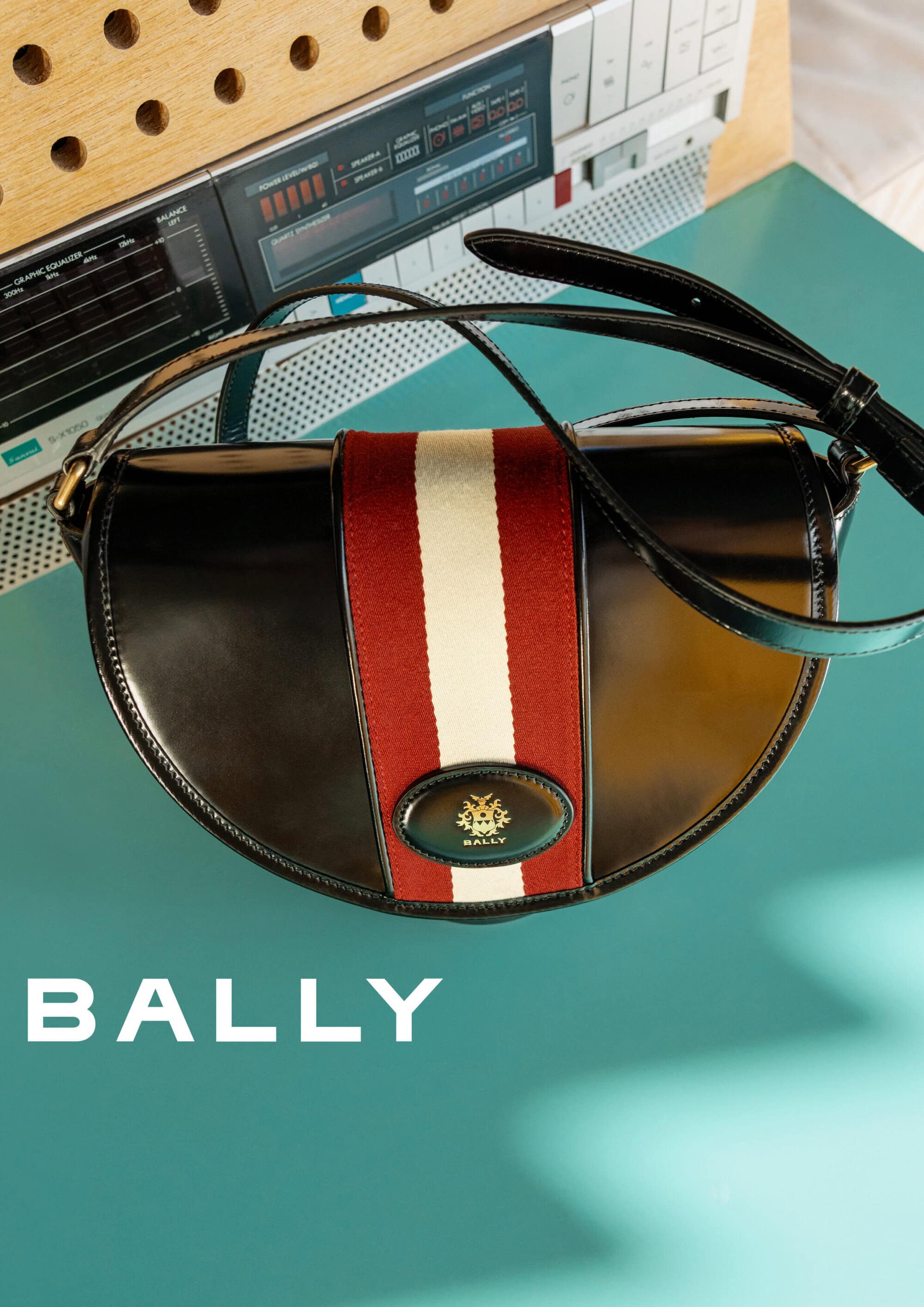 A black leather Bally handbag with a red and white stripe sits on a turquoise surface beneath a vintage audio component.