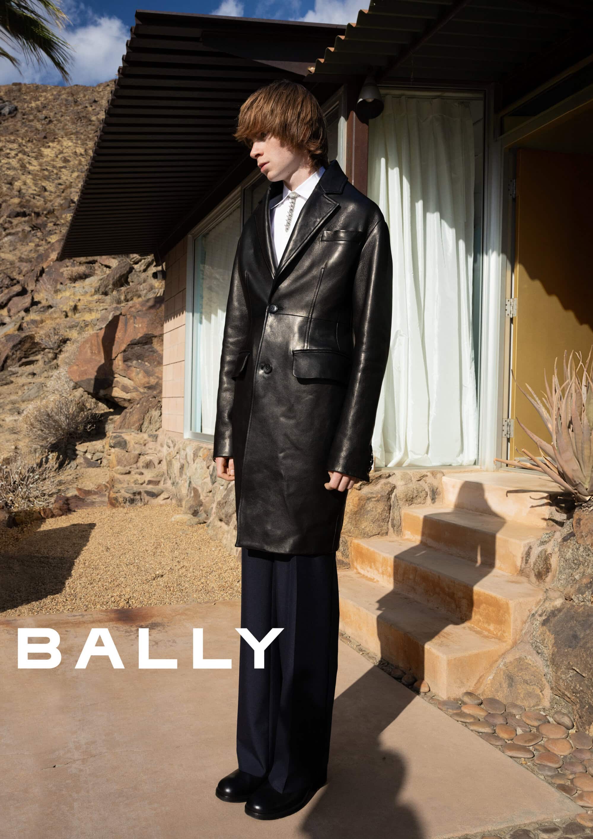 Young man wearing a black leather coat and dark trousers standing outside a modern desert house for Bally Spring 2025 campaign