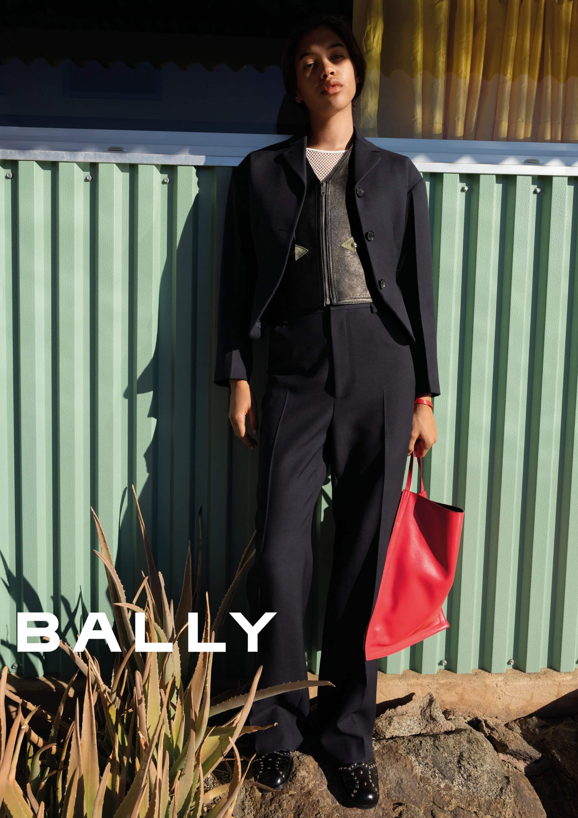 Model standing outdoors wearing a dark suit jacket and trousers holding a red tote bag against a green corrugated wall.