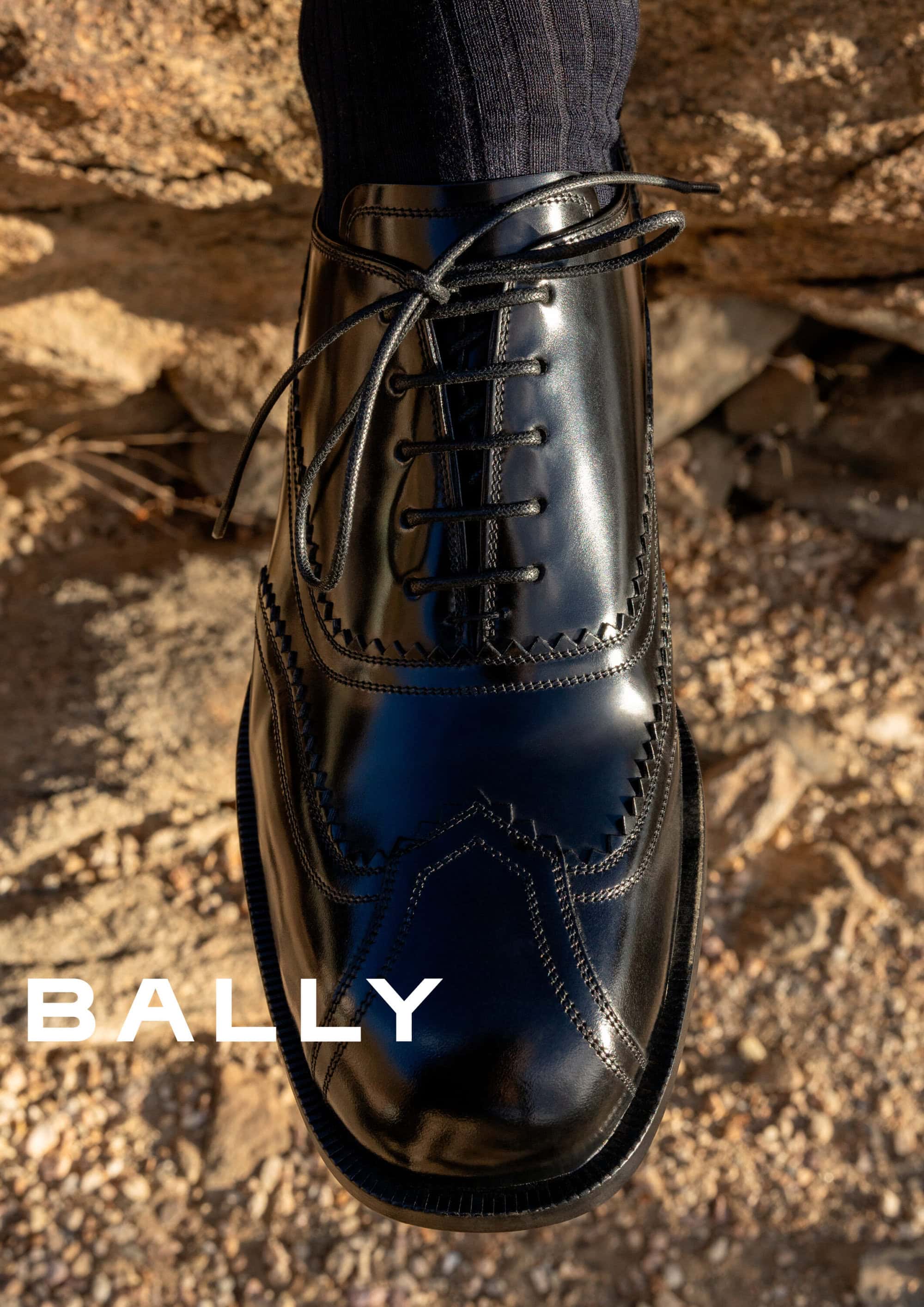 Close-up of a glossy black leather oxford shoe with dark socks against a rocky background for Bally Spring 2025 ad campaign.