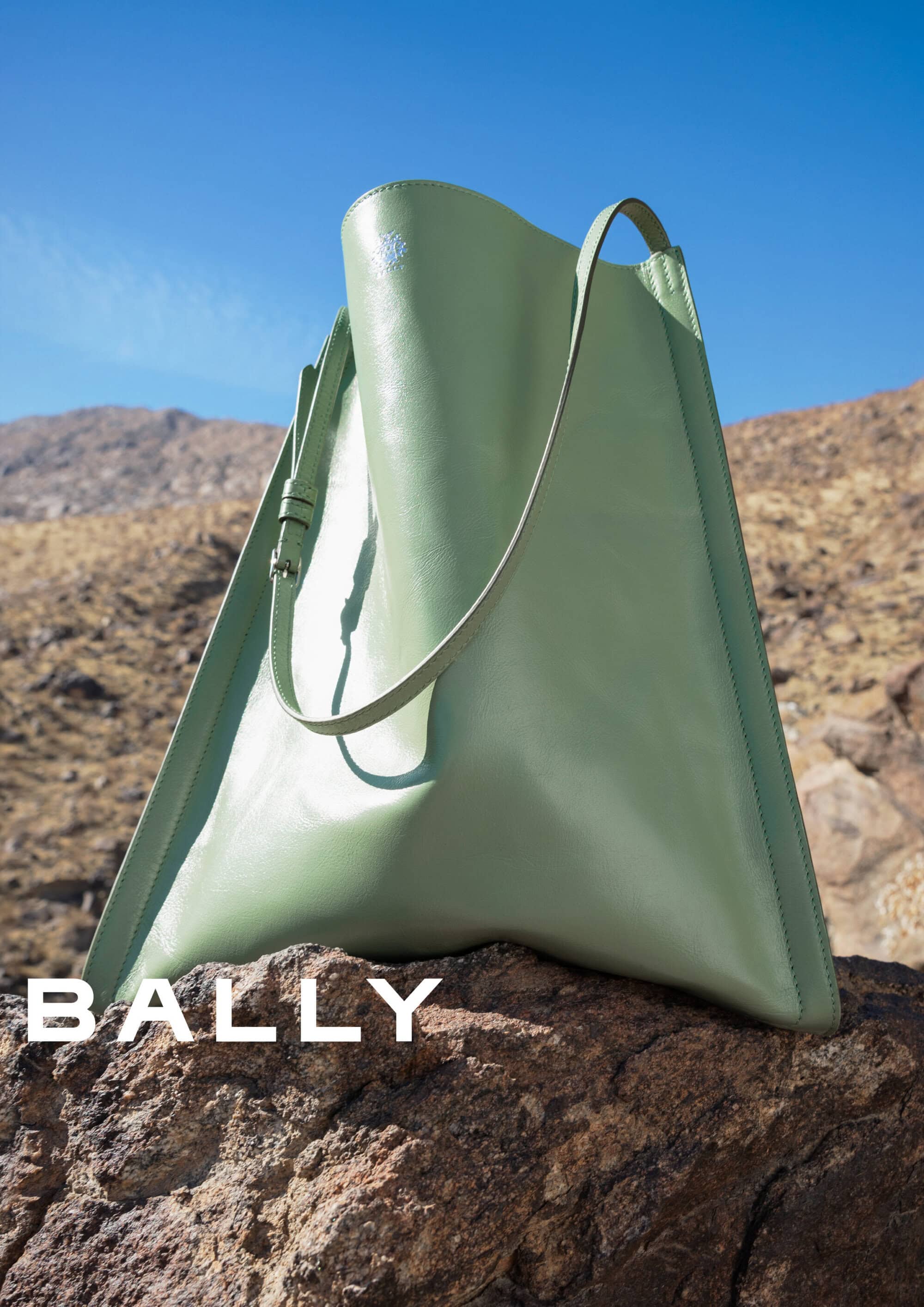 Sage green leather handbag resting on a rugged rock against a dry mountainous landscape and clear blue sky