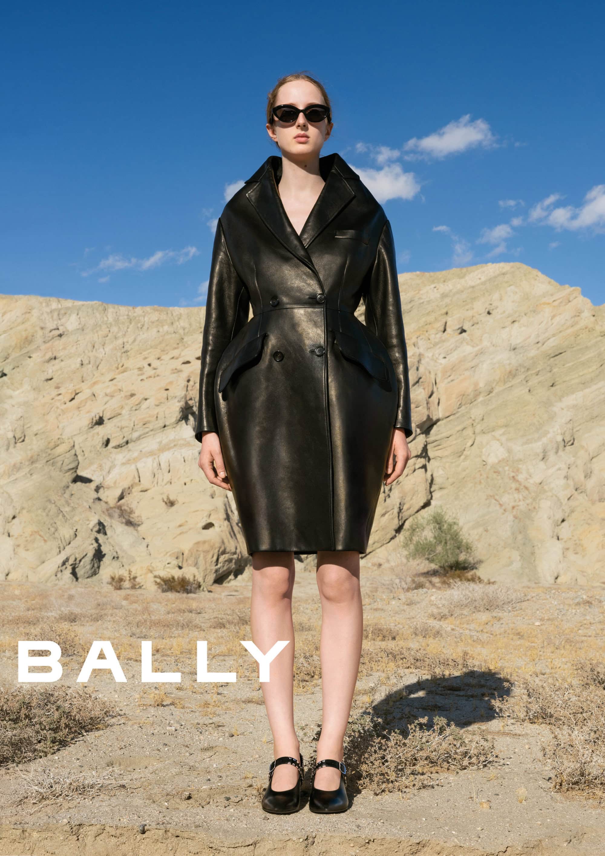Model wearing dark sunglasses and a structured black leather coat standing before a rugged desert landscape under a clear blue sky